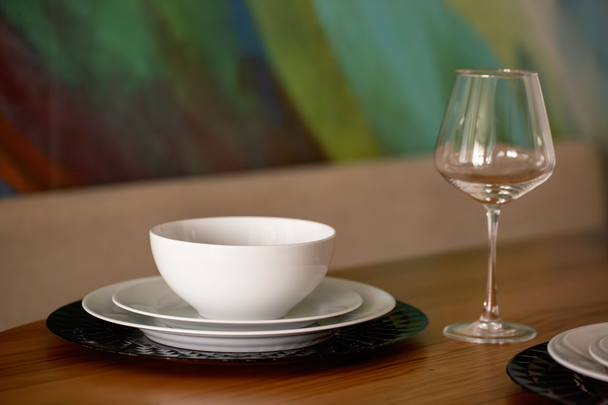 A detailed shot of a dining table setting, showcasing a stack of white dinnerware and a clear wine glass on a wooden surface, with a colorful, abstract painting visible in the soft-focus background.