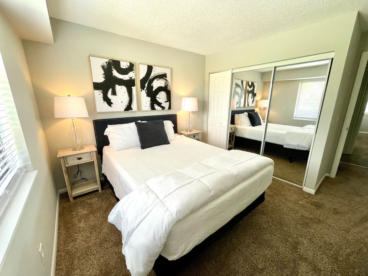Stylish and serene bedroom with plush carpeting, a comfy queen bed, and mirrored closet doors. Thoughtful touches like matching nightstands, modern lamps, and bold black-and-white art make this a restful, inviting space for your short-term stay.