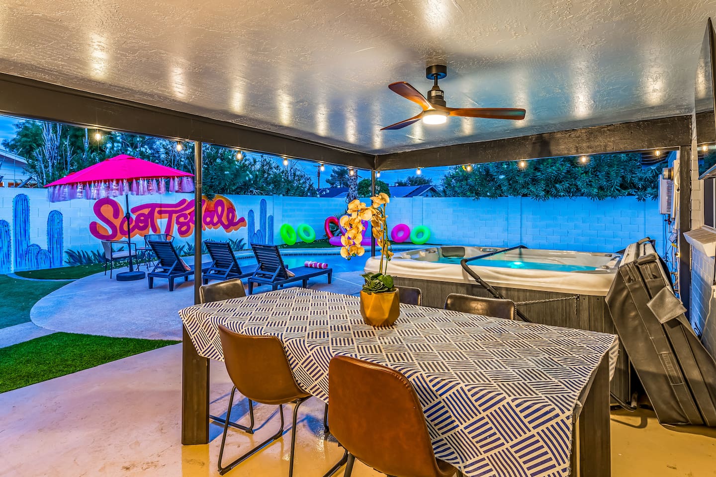 Dine al fresco under the covered patio with string lights, a ceiling fan, and wall-mounted TV—comfort meets open air.

