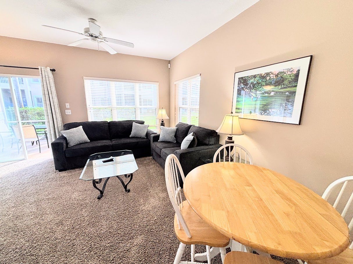 Spacious living and dining area with cozy seating, ceiling fan, round dining table, and natural light from the patio doors.