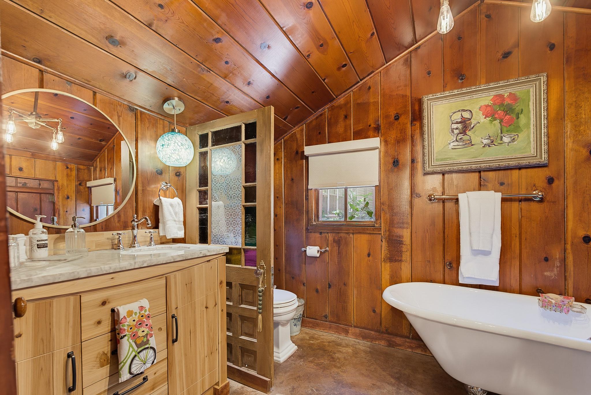 Papaw's Cabin spa-style bathroom with marble vanity, round mirror, stained-glass door, and vintage clawfoot tub. Rustic knotty pine charm meets modern comfort in Waco, Texas.