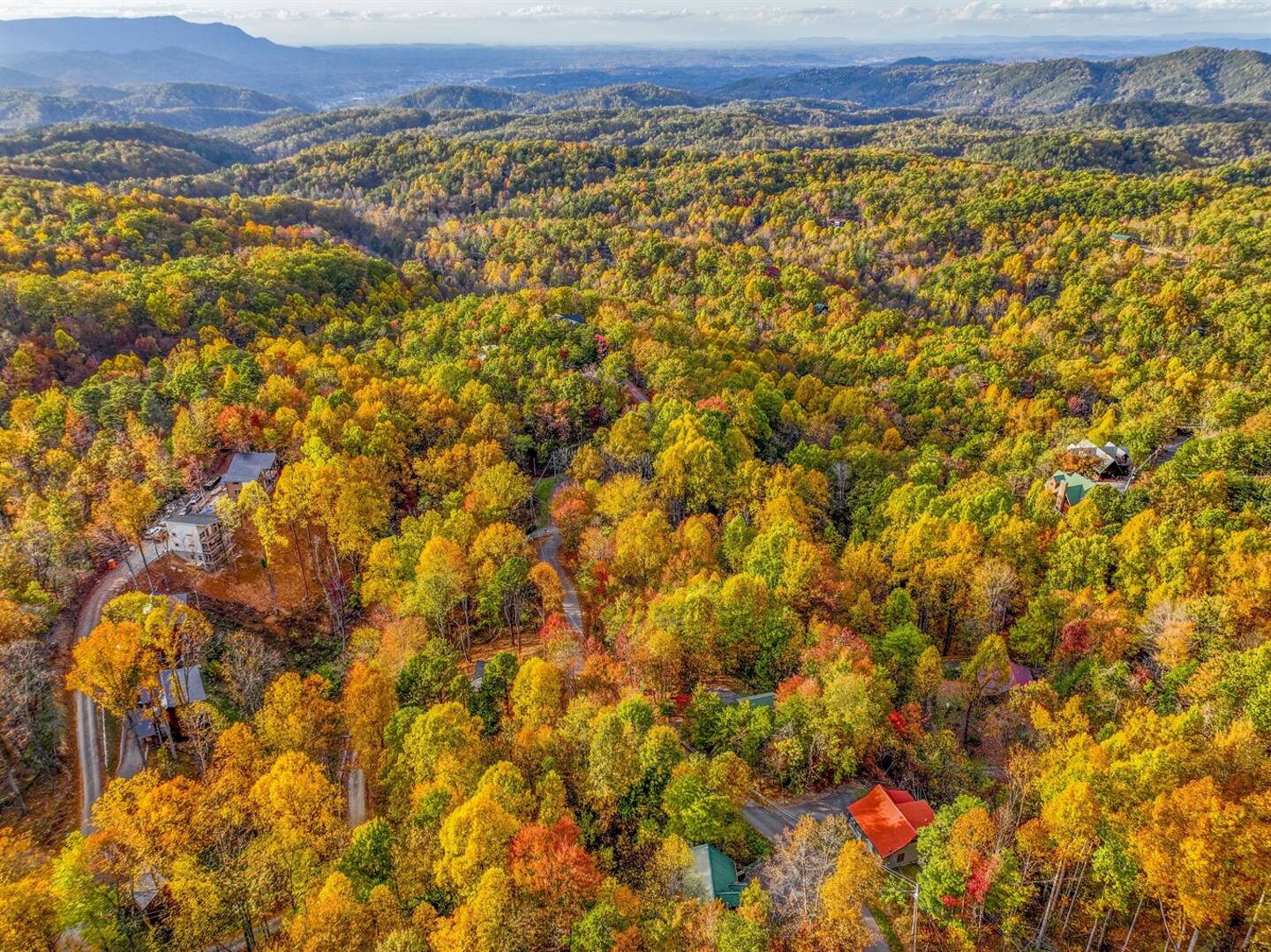 An aerial view of a serene autumn landscape, where vibrant hues of gold, orange, and red blanket the forest. Cozy cabins nestle among the trees, with majestic mountains rising in the distance.