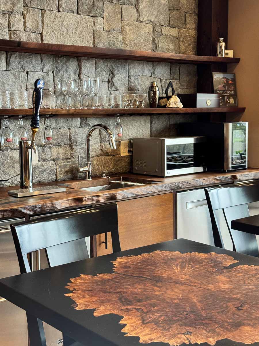 The Tavern Room at Sunset Falls &mdash; live-edge walnut bar, brass tap, and Claro walnut burl epoxy resin dining table on stone wall