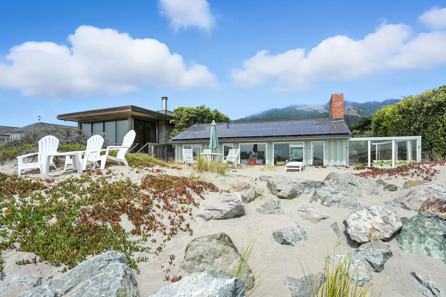Beachfront home with outdoor seating surrounded by sand and rocks.
