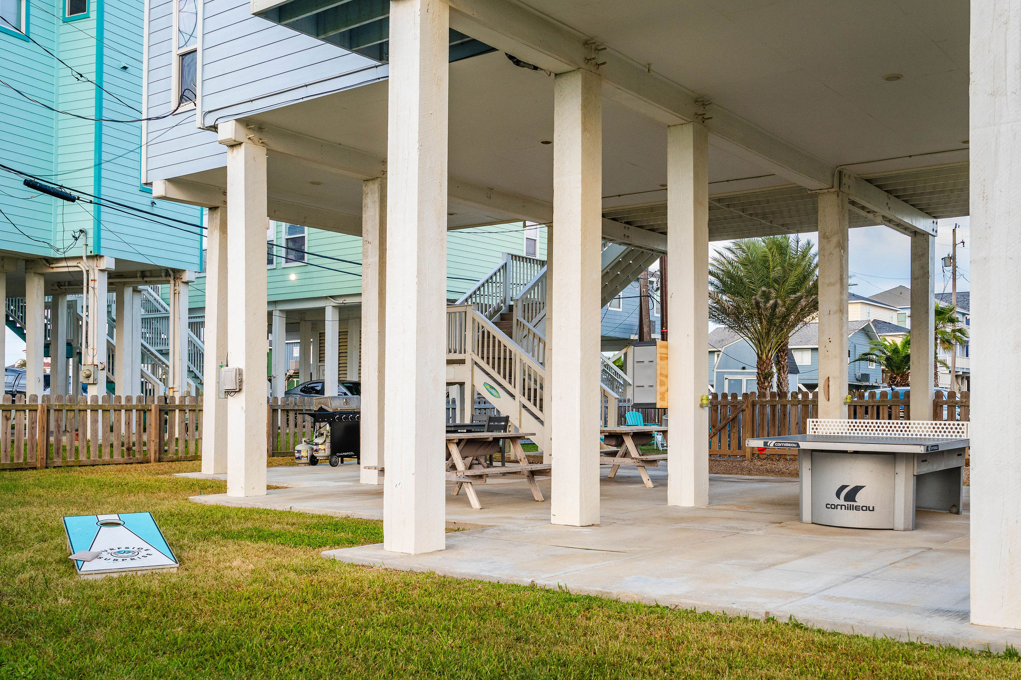 Under-house game zone: cornhole, ping pong, picnic tables, fully shaded. The family tournament writes itself.