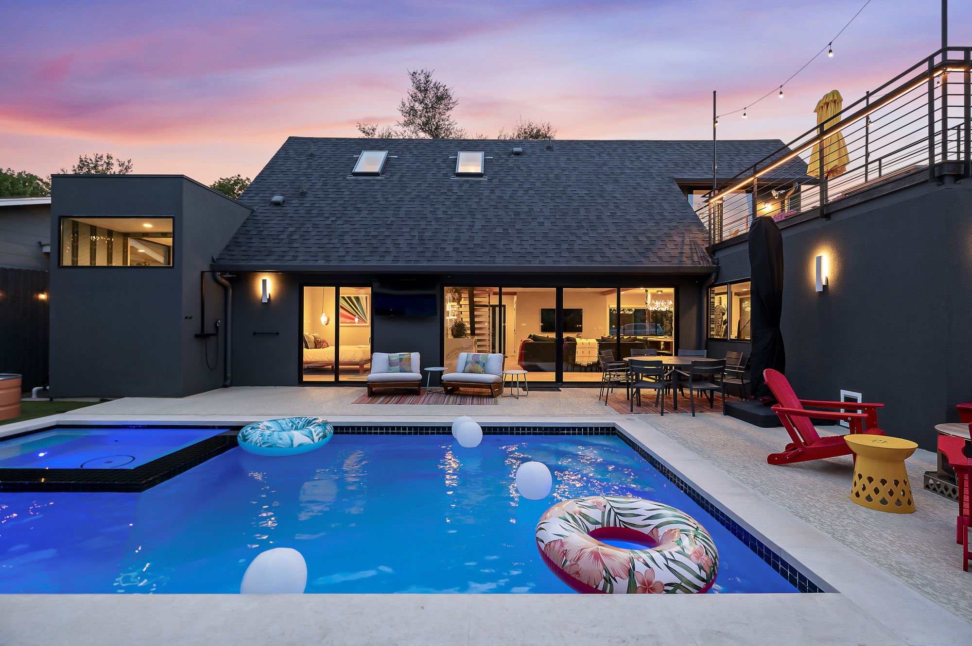 A luxurious backyard with a sparkling blue swimming pool, an adjacent hot tub, and a multi-level deck for dining and lounging, all set against a modern dark gray home with large glass doors and an impressive rooftop balcony.