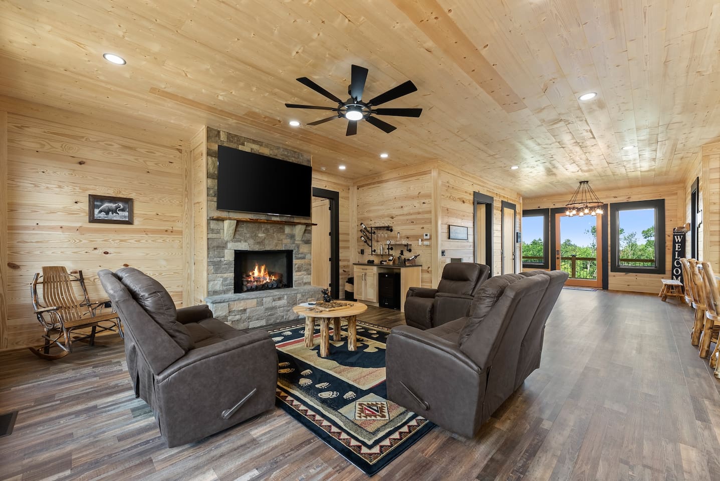  Unwind in the spacious living room featuring plush recliners, a stone fireplace, and a large smart TV—all with serene views of the forest through floor-to-ceiling windows.