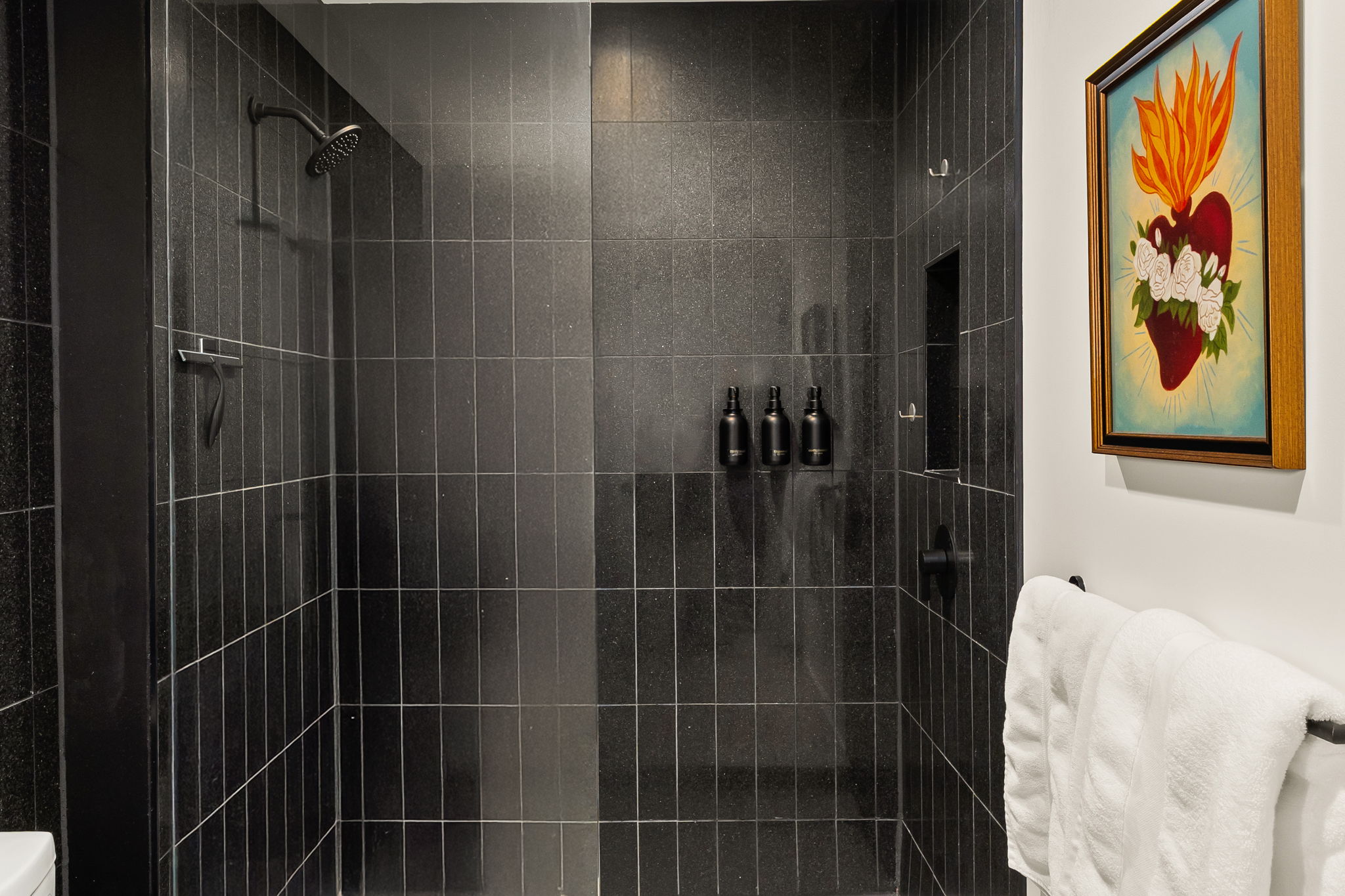 A spacious walk-in shower with sleek black tile, a built-in niche for toiletries, and a modern showerhead, complemented by an artistic, colorful painting of a sacred heart on the wall outside the shower.