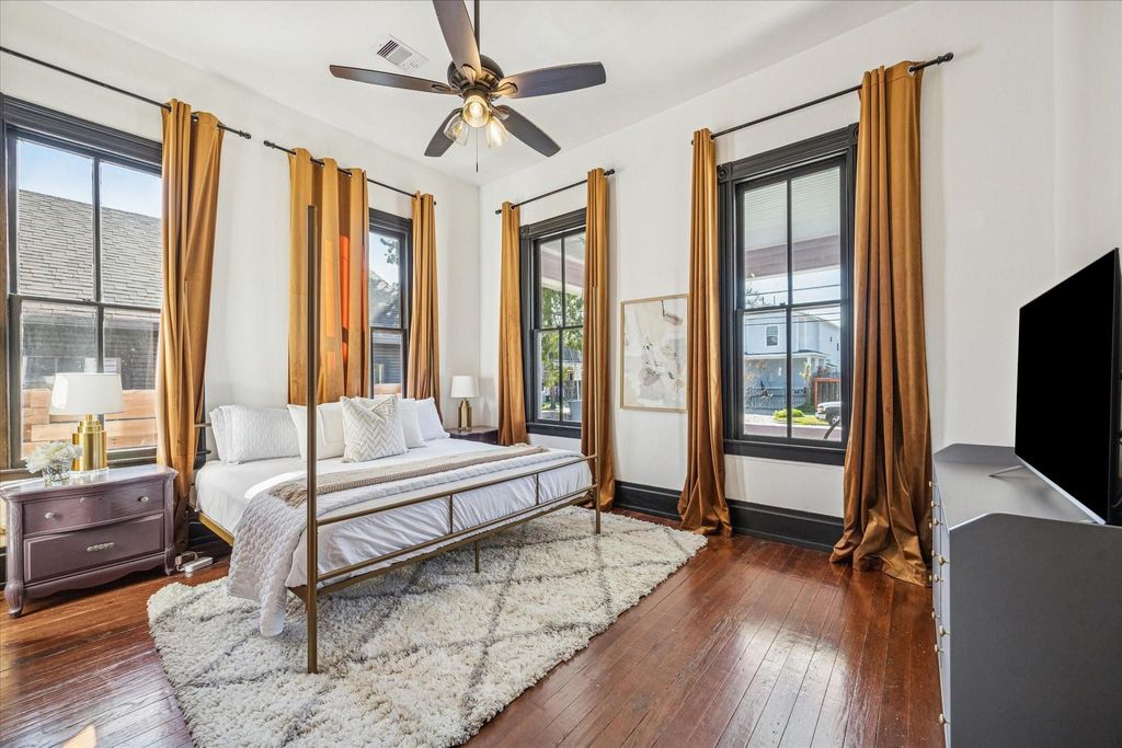 This bedroom blends calm and character with original hardwood floors, tall windows framed by rich millwork, stylish lighting, and a ceiling fan that adds modern comfort to a space steeped in nearly a century of history.