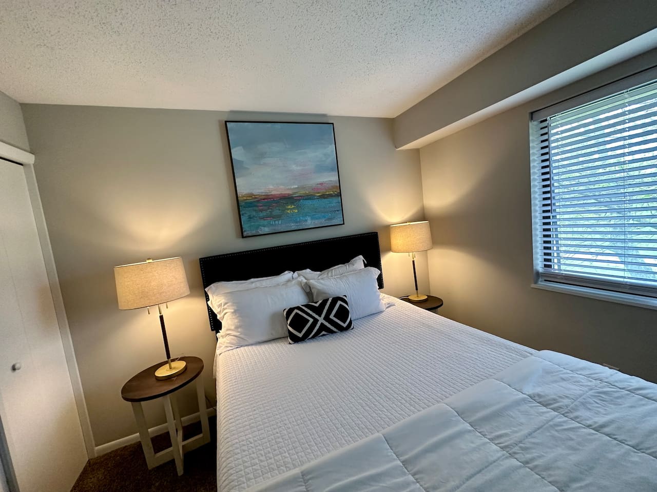 Unwind in this serene bedroom retreat featuring a comfortable queen-size bed, crisp white bedding, and warm accent lighting. A large window fills the room with natural light, while modern decor and blackout shades ensure a peaceful night's rest.