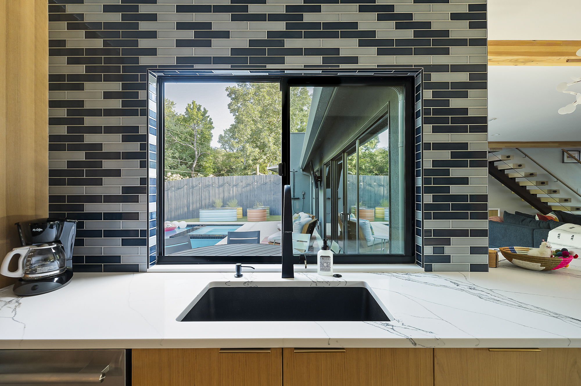 A modern kitchen featuring a black and gray subway tile backsplash, a large window overlooking a backyard with a swimming pool and a deck, and a deep sink with a contemporary black faucet set into a white marble-look countertop.