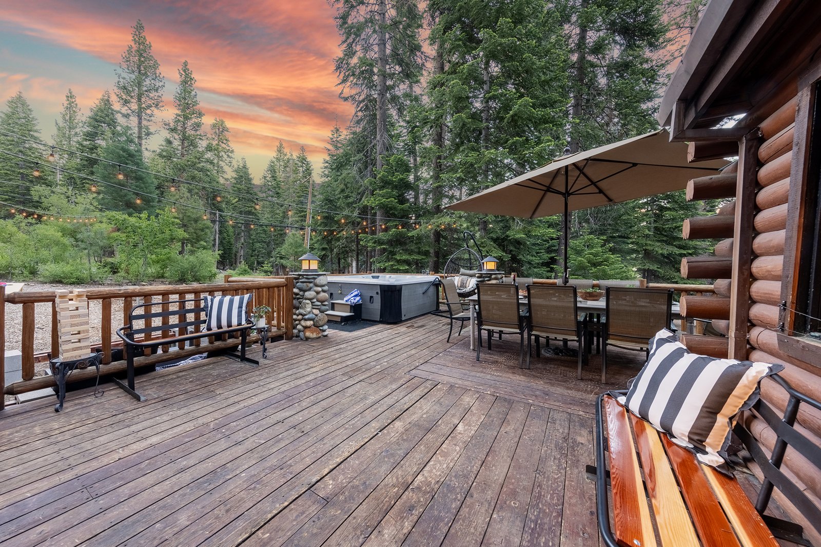 Spread out across the wraparound deck as the sun drops — hot tub on one end, dining in the middle, string lights tying it all together.