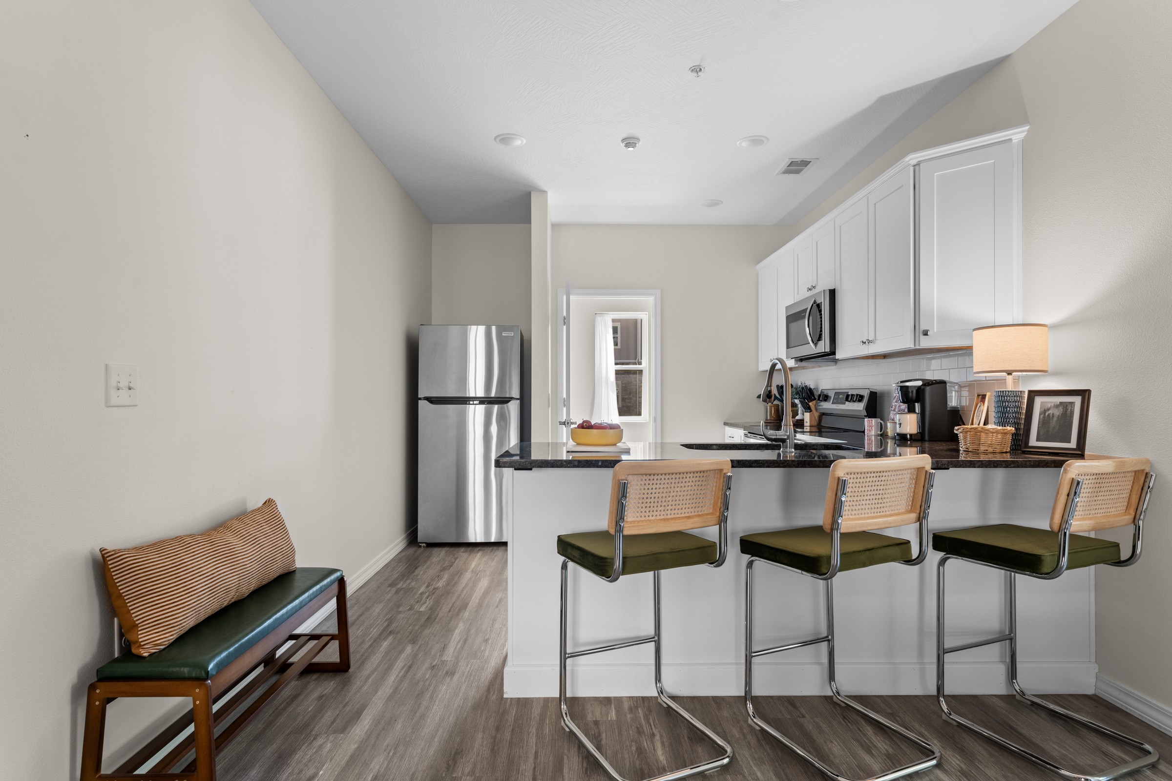 Kitchen peninsula with three cane-back stools and a fridge. A little bench by the entry is handy for tying shoes and dropping bags.