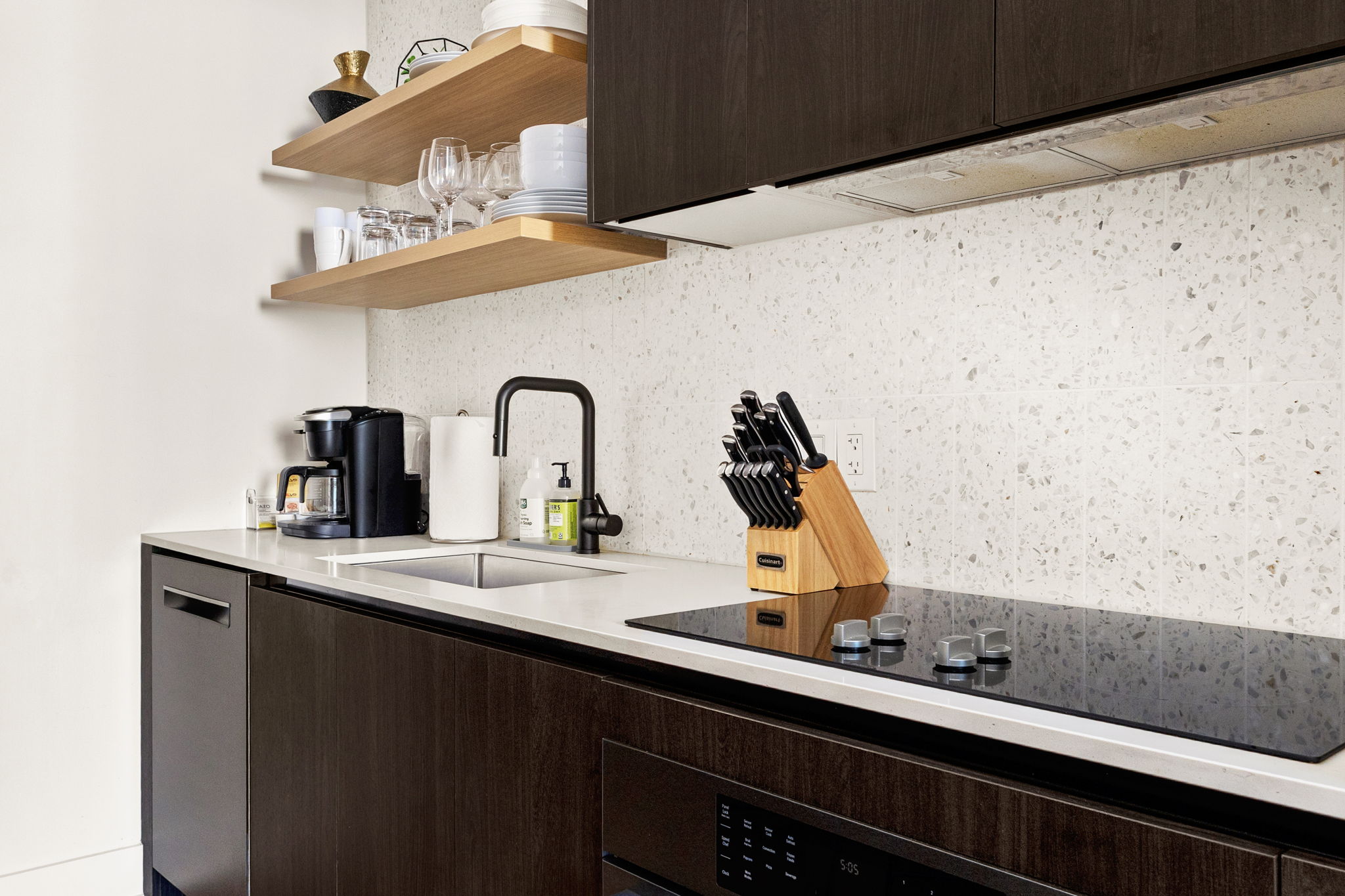 A sleek, modern kitchen with dark wood cabinetry and floating wooden shelves. The space features a light-colored speckled countertop and backsplash, a black gooseneck faucet, a built-in electric stovetop, and a complete knife set, all contributing to a cle