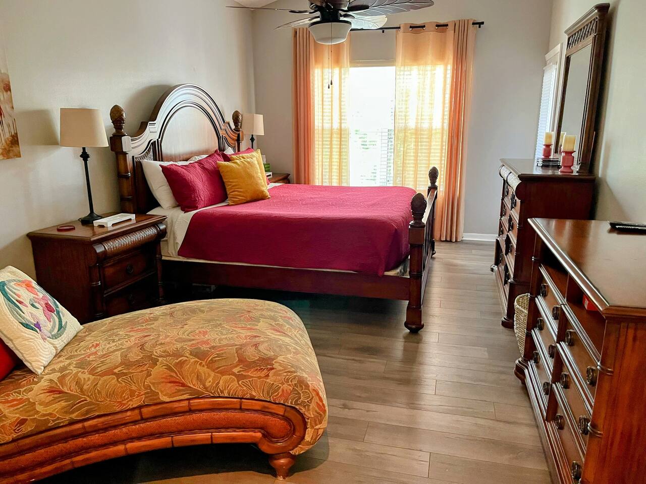 Spacious bedroom featuring a large bed, chaise lounge, wood-look flooring, dresser storage, and warm natural light.