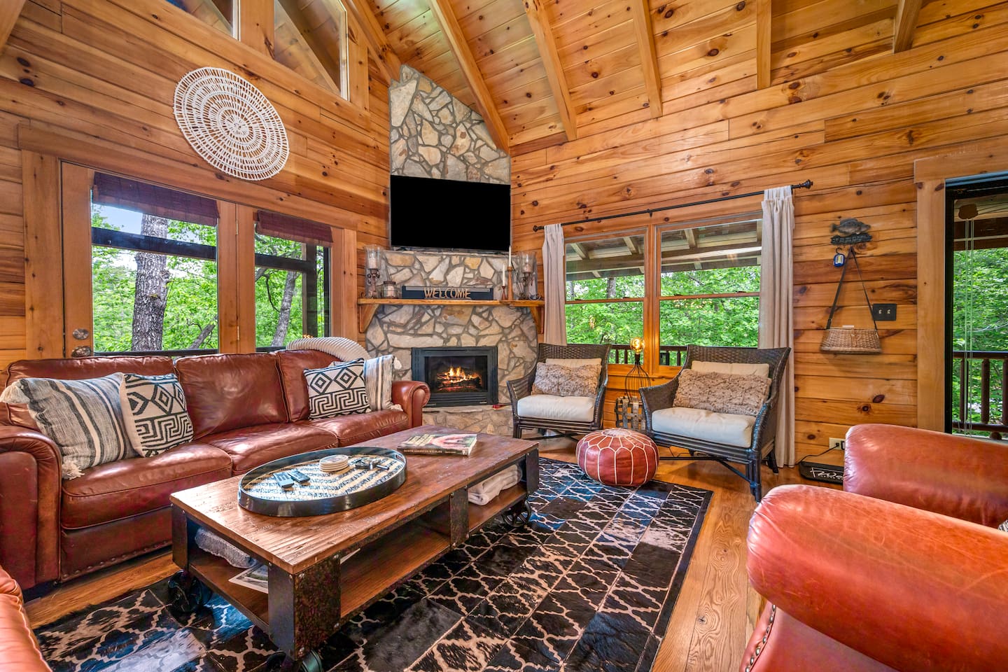 Main living room featuring a beautiful stone fireplace, vaulted wood ceilings, comfortable seating, and large windows overlooking the peaceful wooded surroundings.