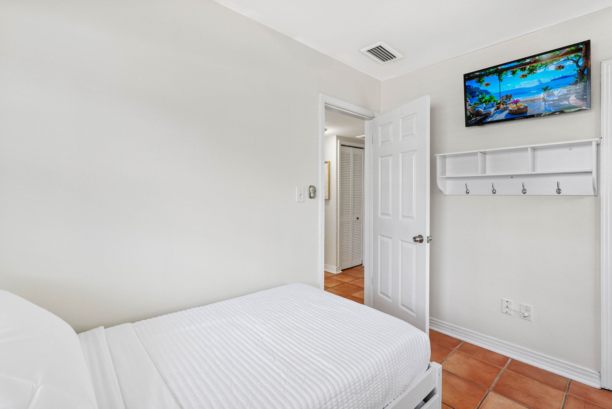 This charming bedroom features a clean, minimalist design with crisp white bedding, terracotta tile floors, and a mounted TV for your entertainment.