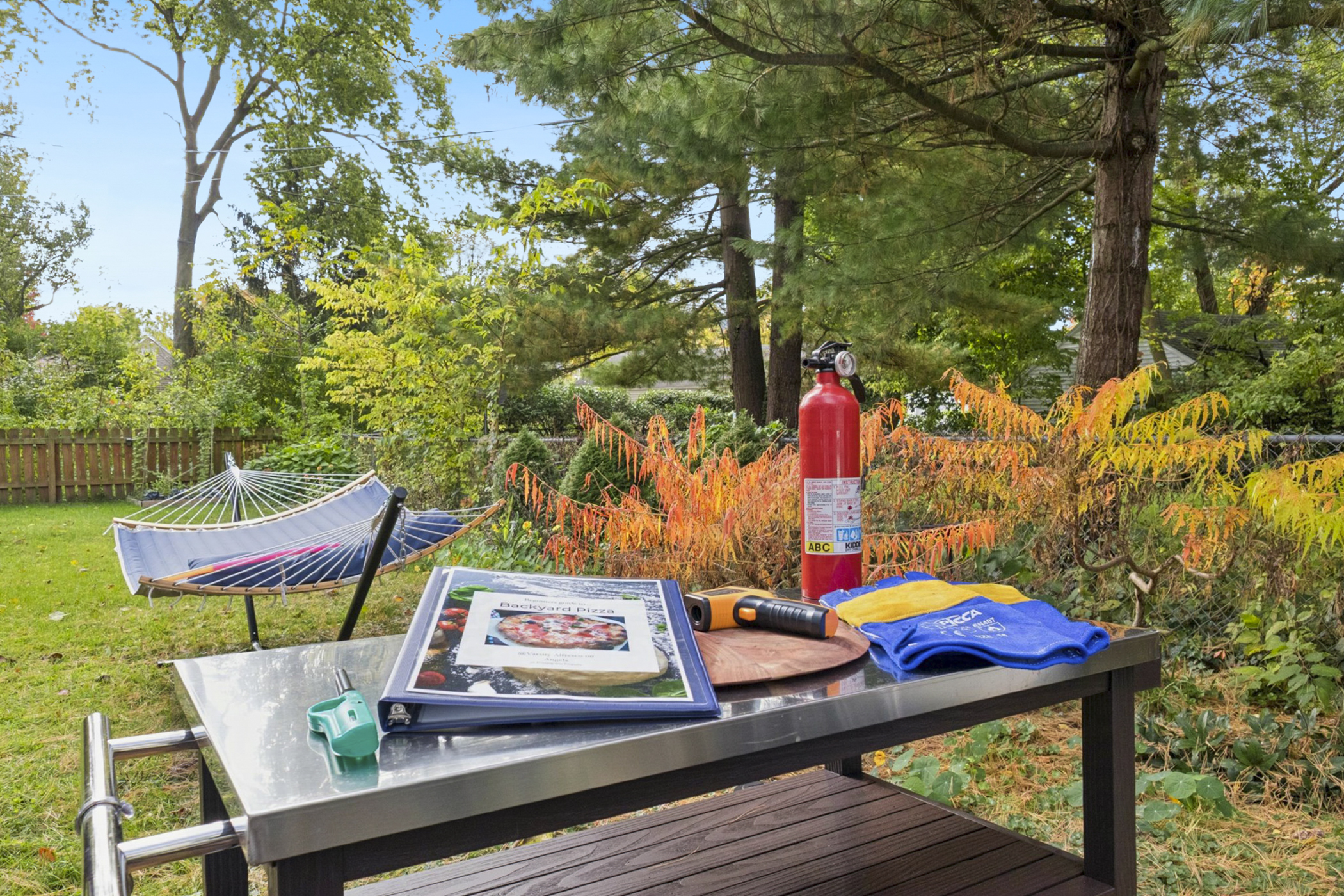 Varsity Pizza Nights are a go—our backyard prep table is stocked with gloves, fire extinguisher, infrared thermometer, lighter, and a pizza guide. Hammock and fenced yard behind complete the scene.