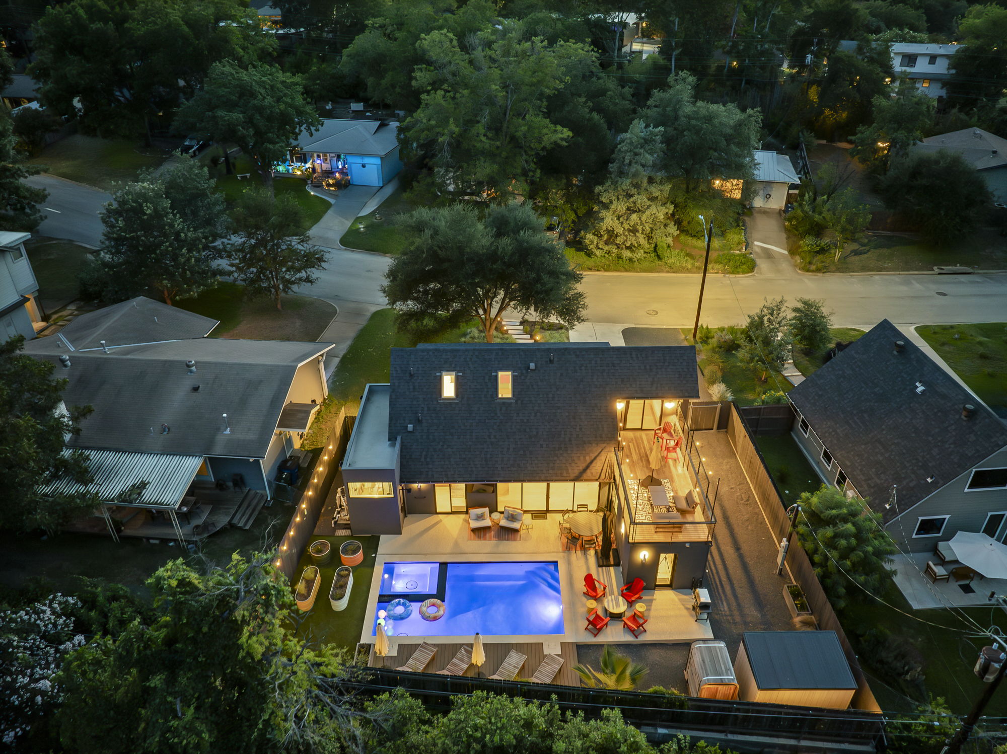An aerial nighttime view of a modern home featuring an illuminated swimming pool with a hot tub and an inviting outdoor entertaining area with a multi-level deck, all nestled within a lush suburban neighborhood.