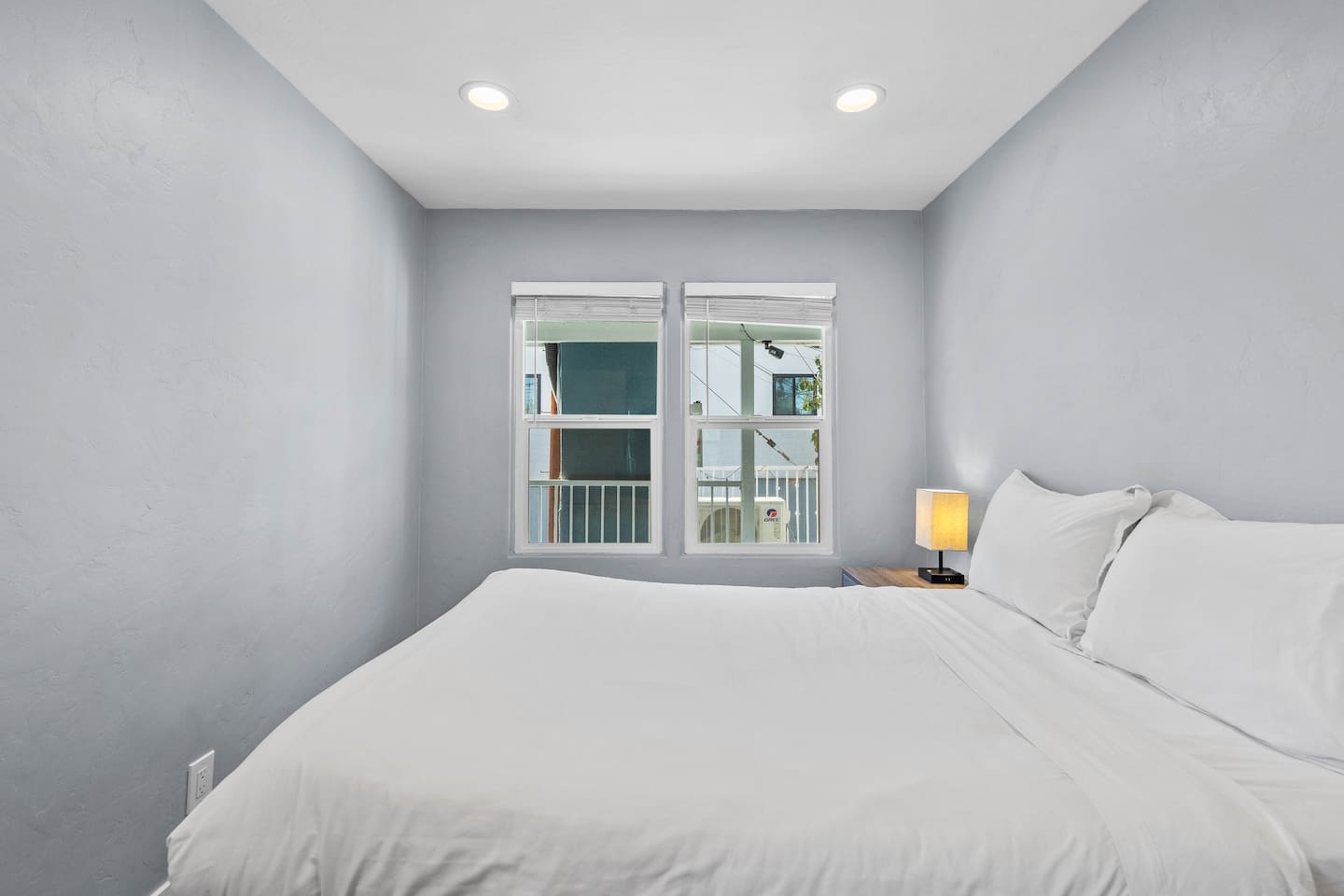 Inviting bedroom featuring a comfortable bed, an aesthetic lampshade providing warm light, and a window with a view of the neighborhood. This cozy setup offers natural light and a peaceful ambiance, perfect for relaxation.