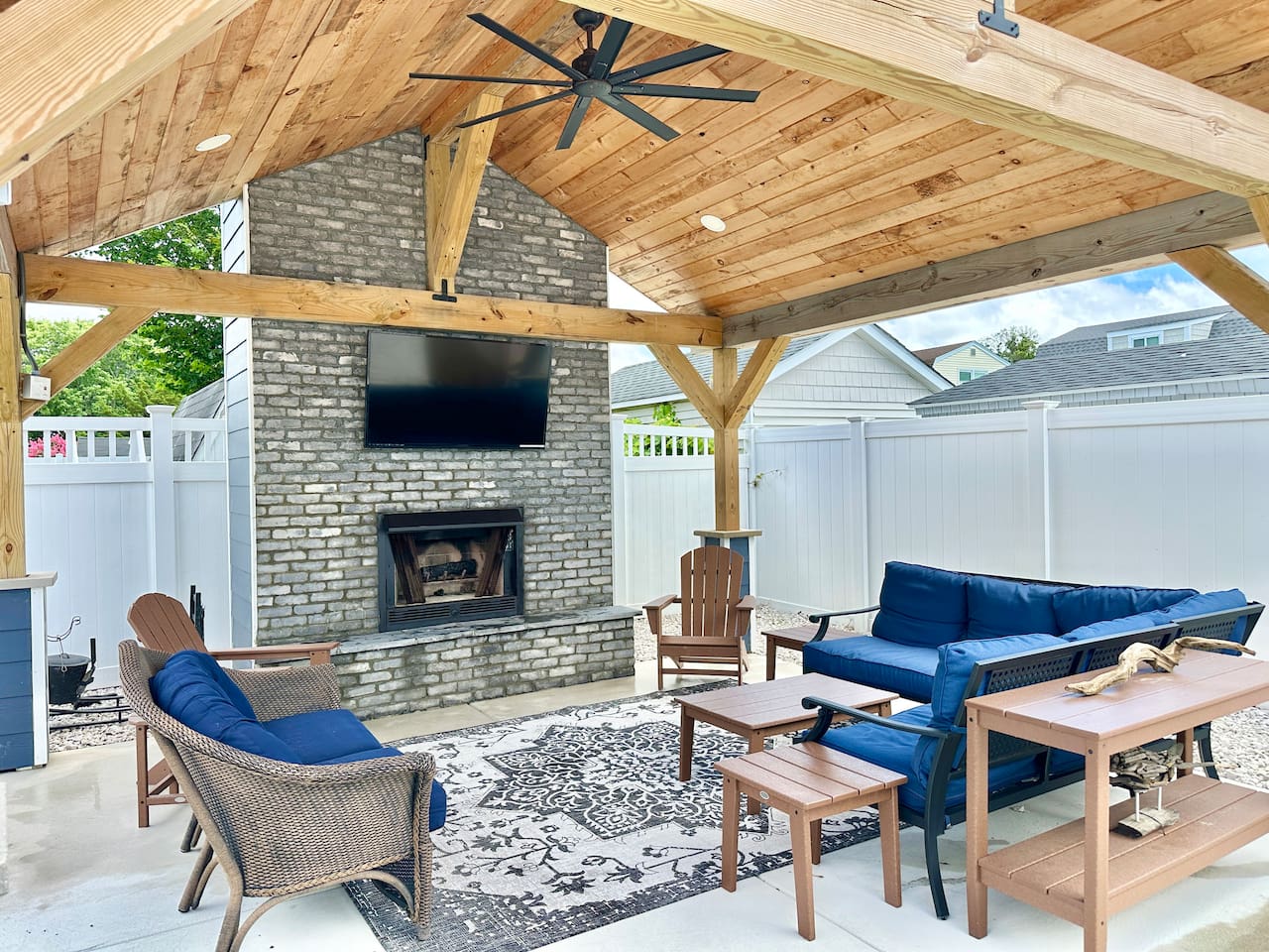 Experience luxury outdoor living in this stunning covered patio! Featuring a rustic wood ceiling, a grand stone fireplace with a TV, and plush blue seating, it’s the perfect spot for breezy summer evenings or cozy fall nights.