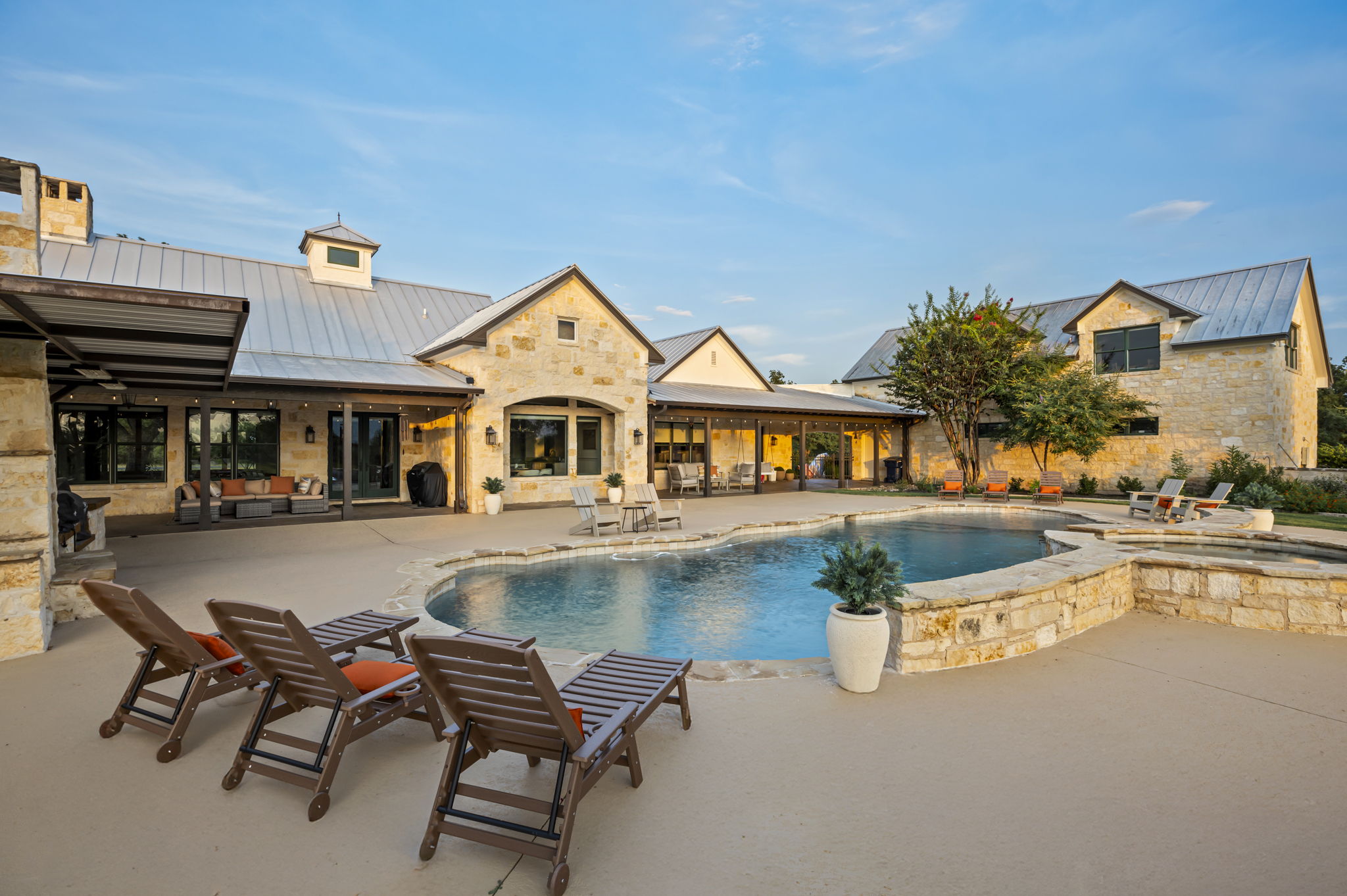 A home featuring a sprawling outdoor living area with a resort-style pool, hot tub, and ample lounging space, set against a clear blue sky.