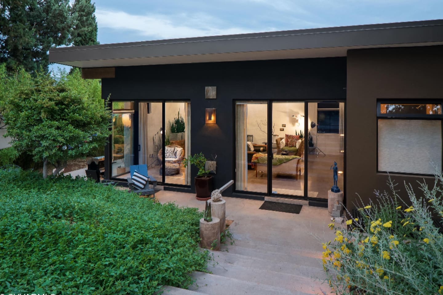 Modern retreat at dusk with warm interior glow, expansive glass doors, and serene garden surroundings.