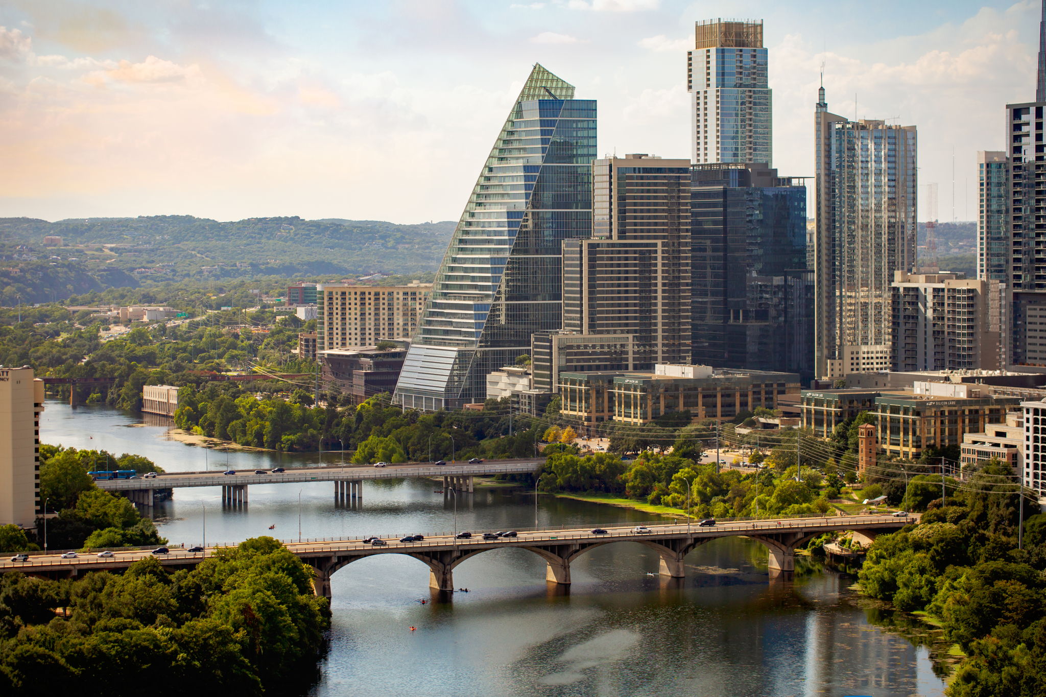 A vibrant aerial view of a bustling city skyline with modern skyscrapers, a river flowing through the urban landscape, and lush green trees lining the banks and hillsides in the background.