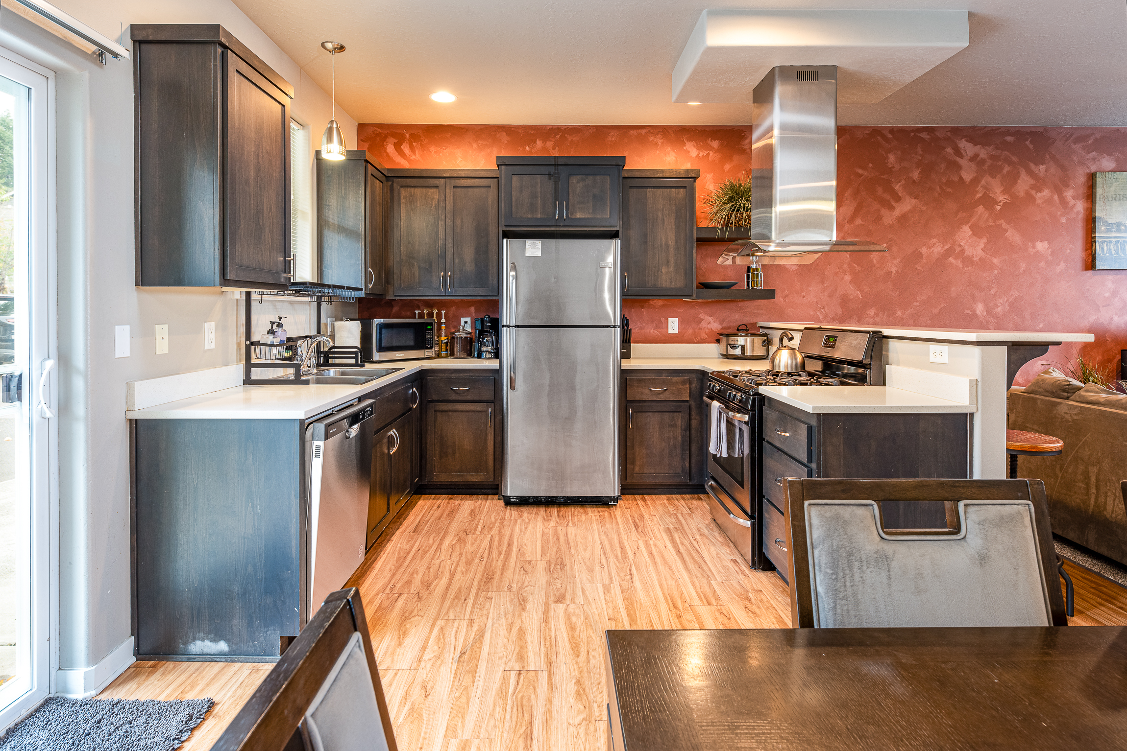 A modern kitchen featuring dark wood cabinets, stainless steel appliances including a refrigerator and stove, a light-colored countertop, pendant lighting, and a reddish-orange textured wall, with a dining table and chair visible in the foreground.