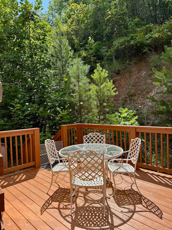Cozy deck with glass table & white chairs. Lush greenery.