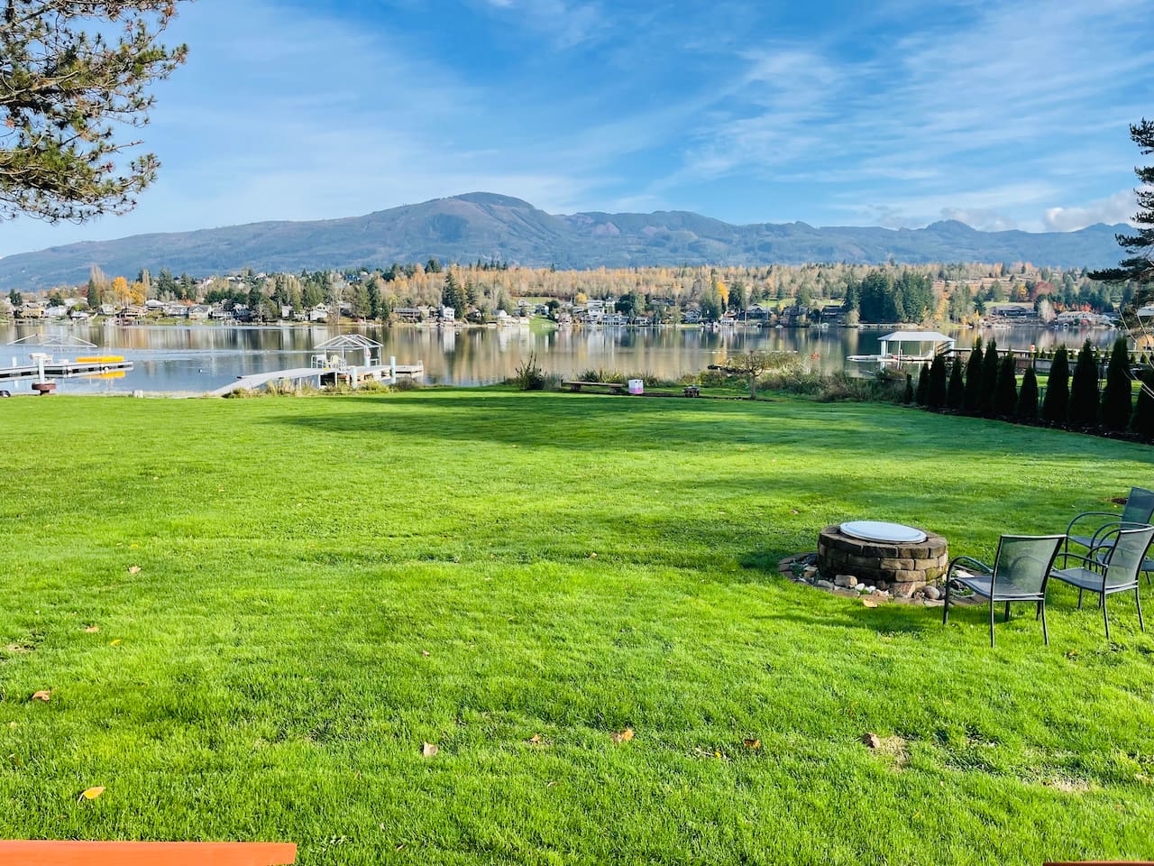 A wide, green backyard stretches to the lake, with a dock and fire pit ready for evenings outdoors. The open lawn feels inviting, and the calm water reflects the shoreline, creating a peaceful connection between land and lake.