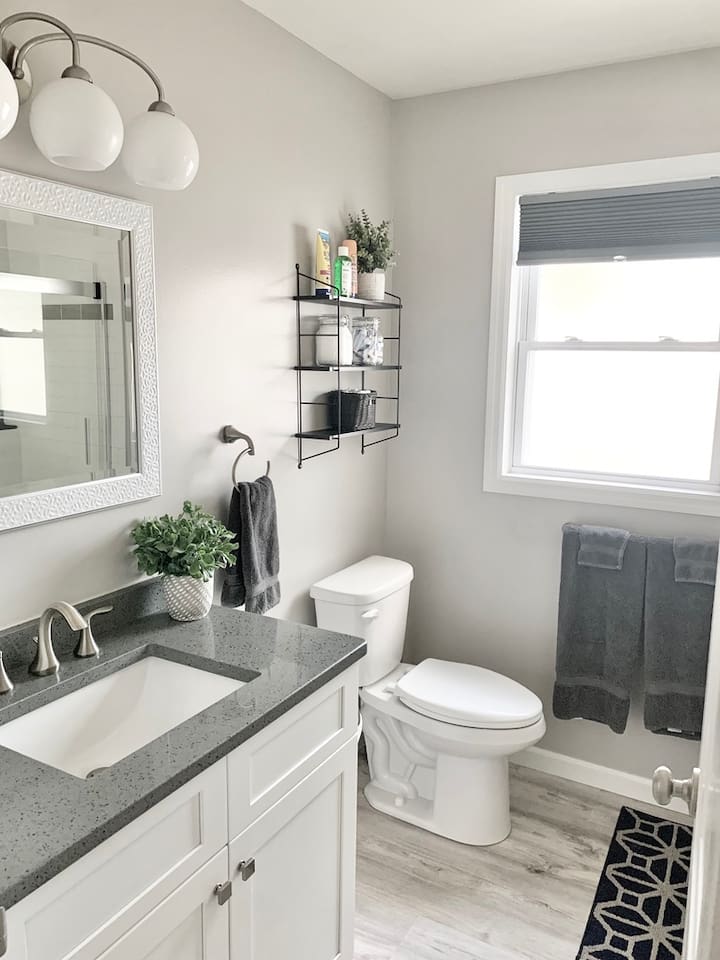 Refresh in this bright, updated bathroom featuring a sleek grey quartz vanity top and a white shaker-style cabinet. The space is finished with clean white walls, dark charcoal towels, and a window that provides plenty of natural light.