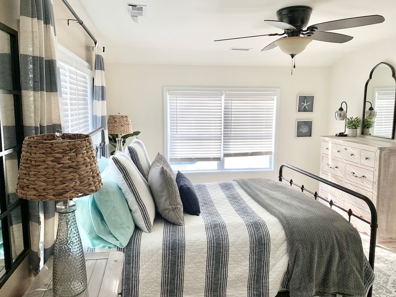 Wake up in a sun-drenched sanctuary featuring a black iron bed frame and plush nautical-themed bedding. This bright primary bedroom is complete with rustic wood furniture, woven textures, and soft coastal accents for a truly peaceful stay.