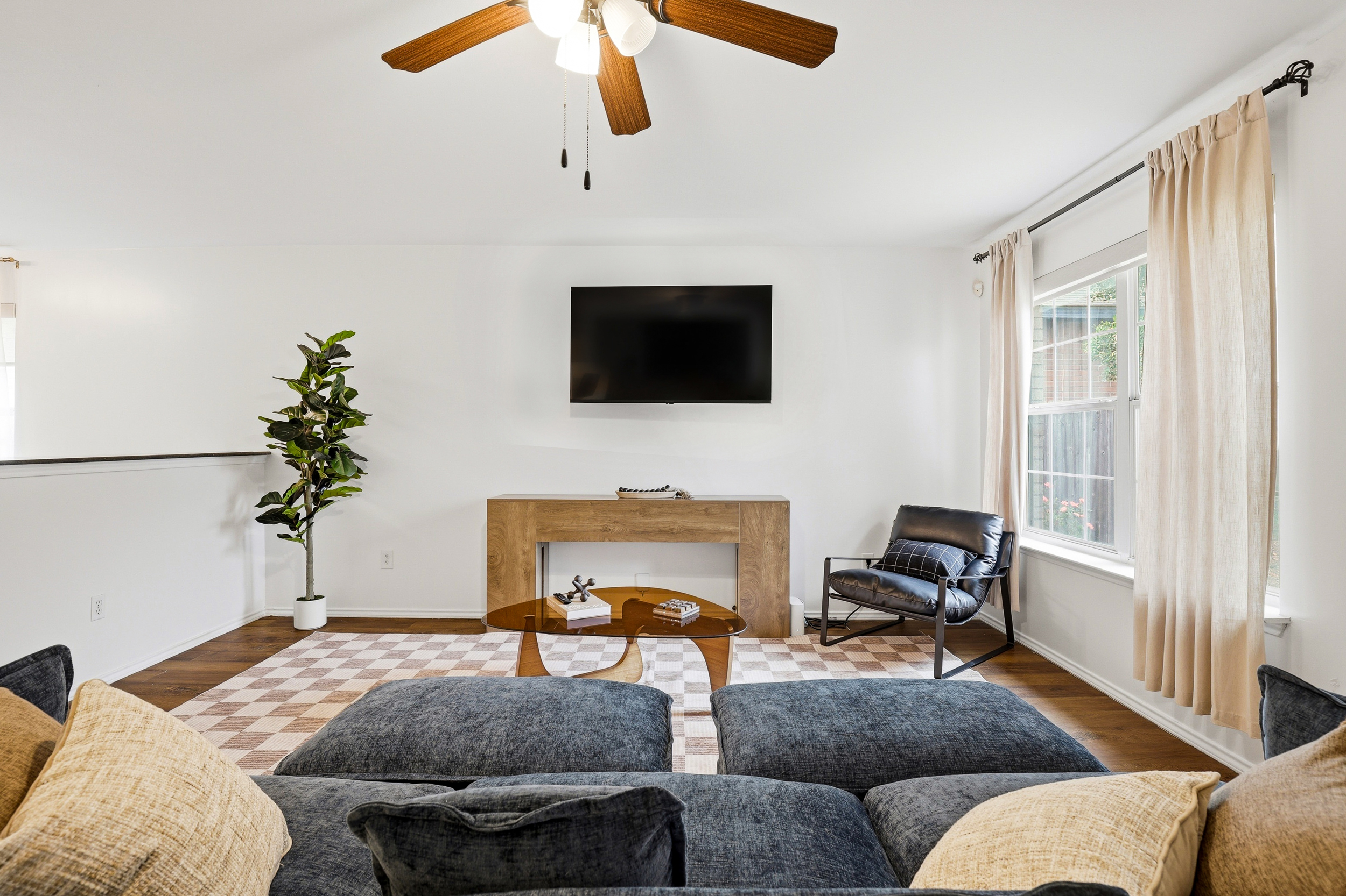 Wall-mounted Smart TV, wood console, accent chair, and ceiling fan in the main living area. Bright, clean space designed for comfort with room-darkening curtains for movie nights.