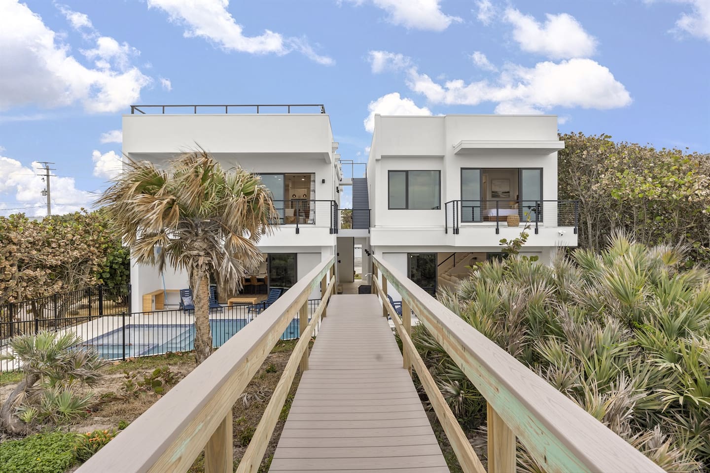 Admire the exterior of the house with lush trees surrounding a sparkling swimming pool, a spacious balcony, and a glass door offering access to the indoors, all leading toward a charming bridge, captured from a unique angle