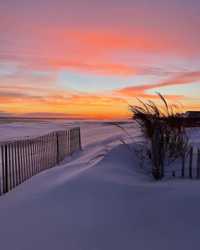 Experience the quiet beauty of the coast in winter. Snow-covered dunes and a rustic sand fence lead toward the ocean, all illuminated by a vibrant sunset of pink and orange.