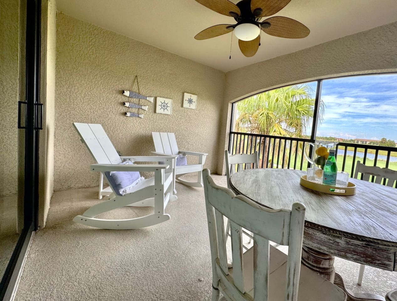 Cozy screened lanai with rocking chairs, round table with 4 chairs. The view outside shows palm trees, a golf course, and a lake.
