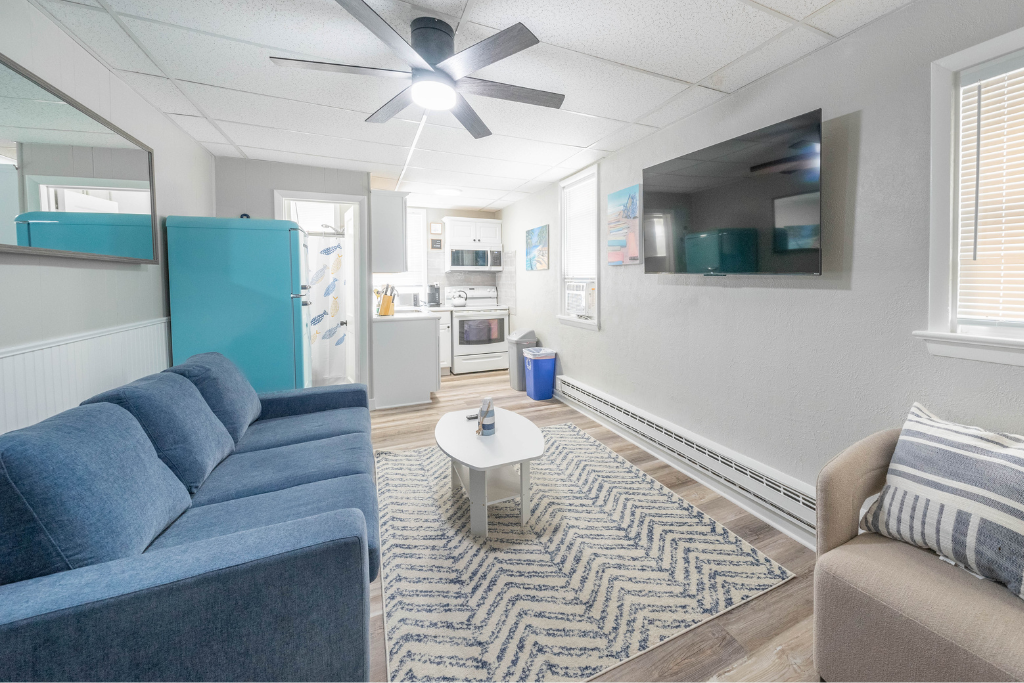 Bright open living area with comfortable seating, ceiling fan, and wall-mounted TV. The layout flows easily into the kitchen, creating a relaxed space for watching shows, unwinding after the beach, or spending time together indoors.