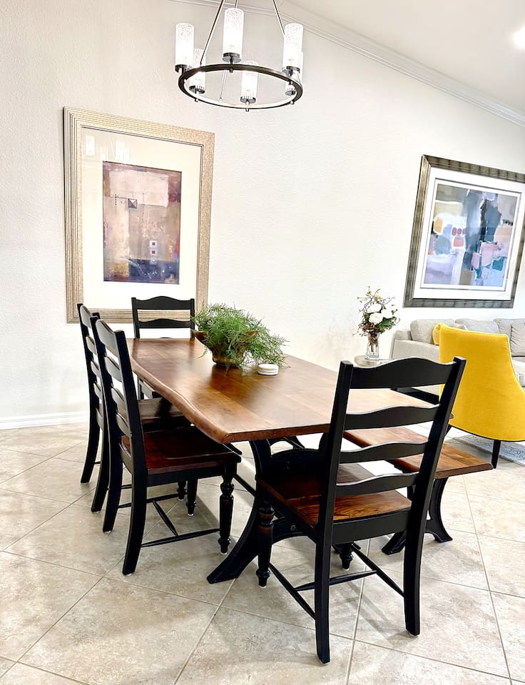 A Daning area with a Acacia wood table, 4 chairs and one bench that can seat up to 7 people. Abstract painting on the wall, light-Colored tiles and Off-white walls.