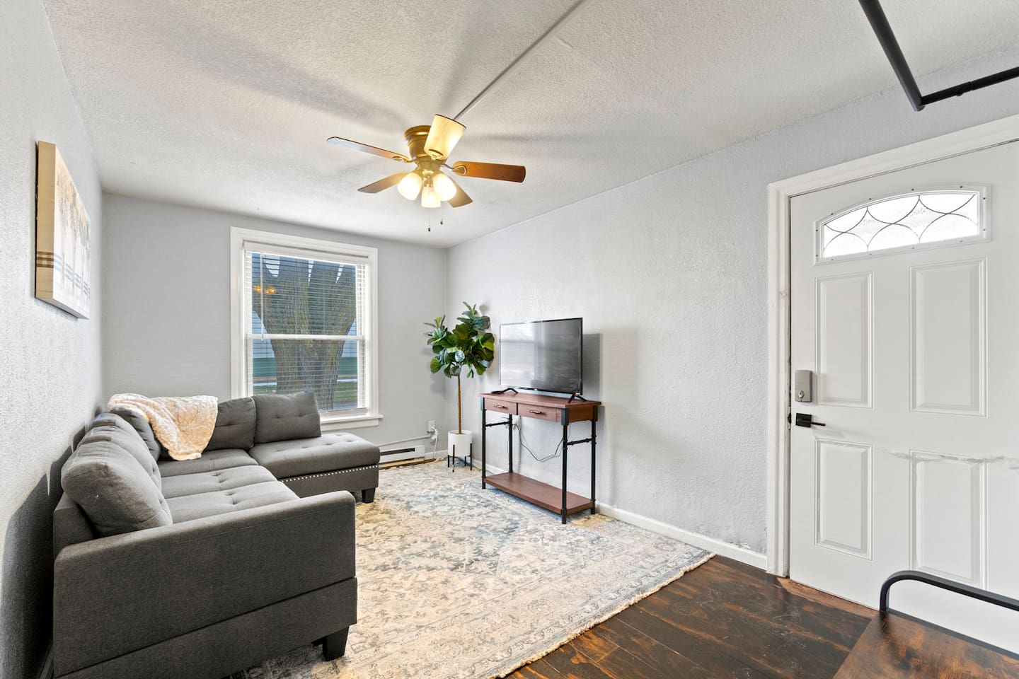 Inviting living room with a bright window, featuring a grey sectional couch with a cozy throw blanket and a TV for ultimate comfort and relaxation."