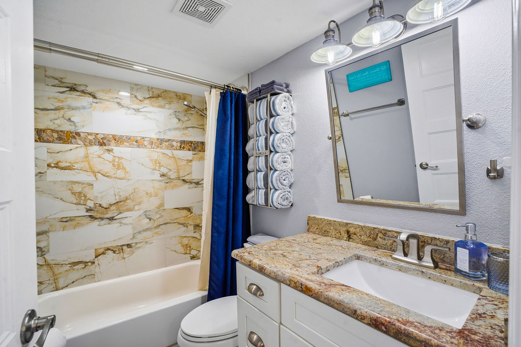 Clean full bathroom featuring a marble tile shower and tub combo, granite vanity countertop, fresh towels, and modern lighting—perfect for a comfortable and relaxing stay near the beach.