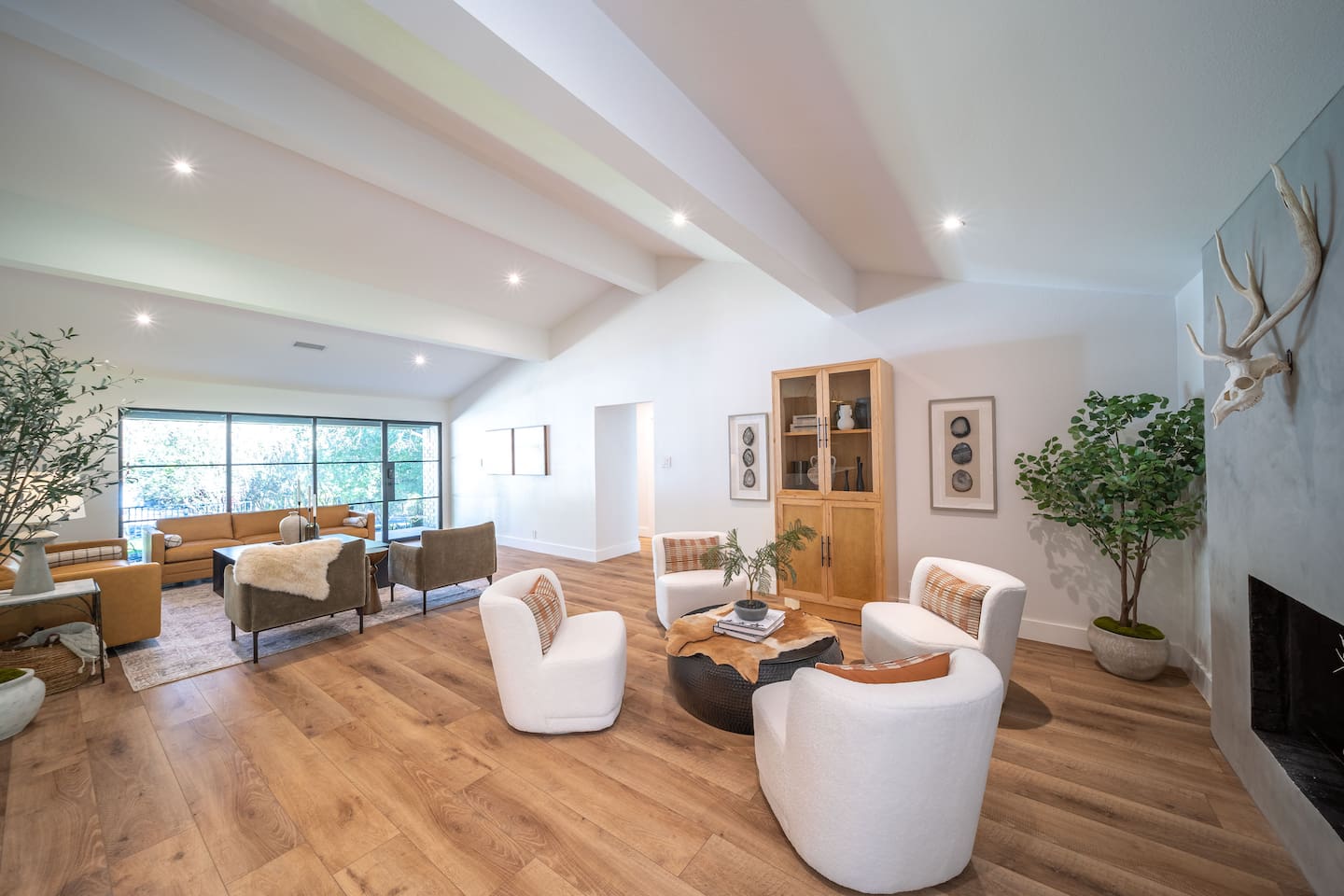 Open-concept living room with designer furnishings, plush seating, and warm textures. Large windows and glass door fill the space with natural light, creating a comfortable gathering area ideal for relaxing or entertaining.