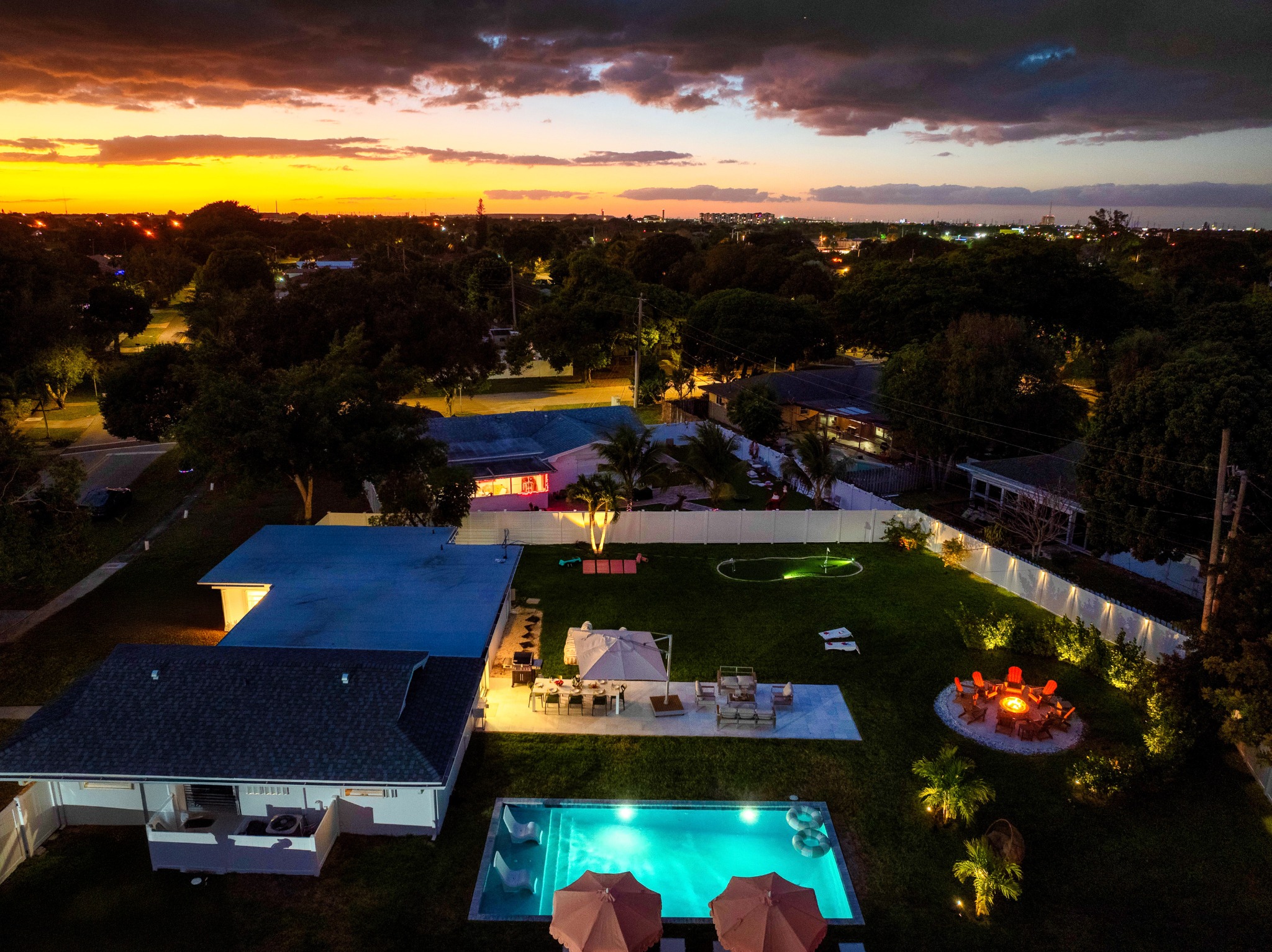 Private Palm Beach backyard at sunset with illuminated pool, fire pit seating, and landscaped grounds.