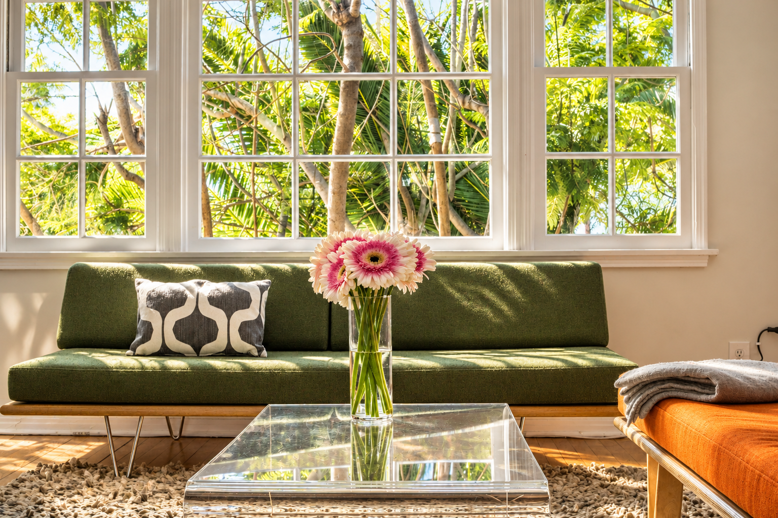 Sunlight pours through the living room windows, with leafy greenery outside and a green upholstered sofa below. Pink flowers, a clear acrylic coffee table and a glimpse of the orange sofa bring bright color and mid-century character to the seating area.