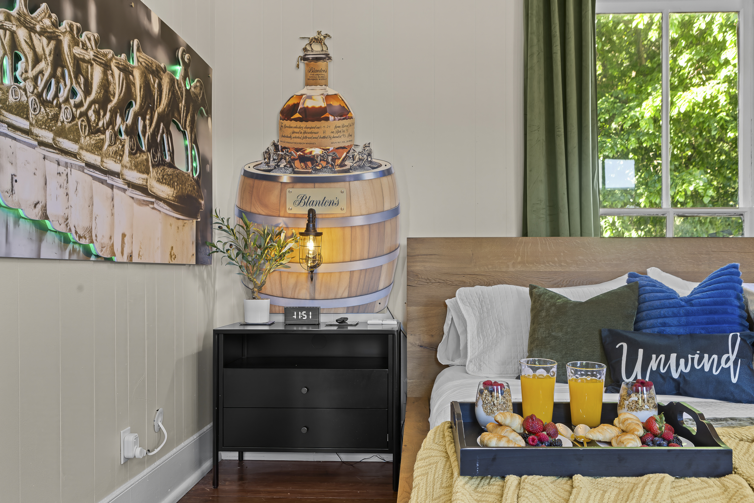 Green curtains, a Blanton’s barrel lamp, and bedside reading within reach make for a well-considered nightstand in a thoughtfully designed room.
