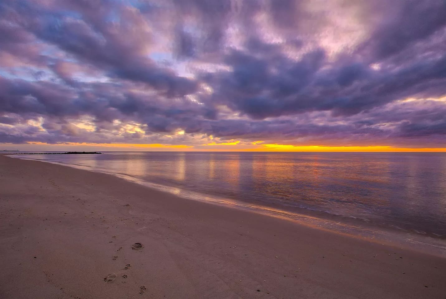 Experience the beauty of a coastal twilight. The sky transforms into a stunning canvas of deep purples and oranges, reflecting over the calm waters and quiet shoreline for a peaceful end to your day.