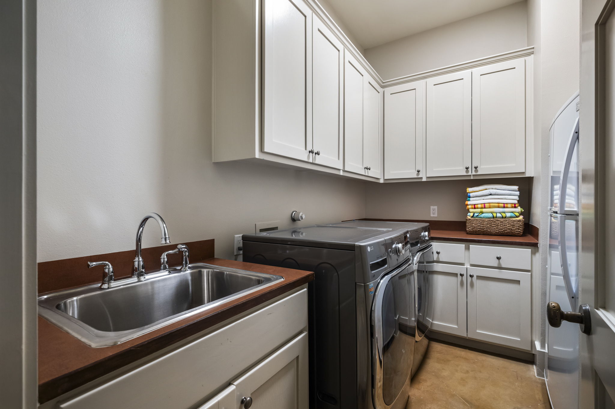 A well-equipped laundry room featuring a stainless steel utility sink, modern front-loading washer and dryer, white cabinetry for storage, and a countertop with stacked, colorful towels.