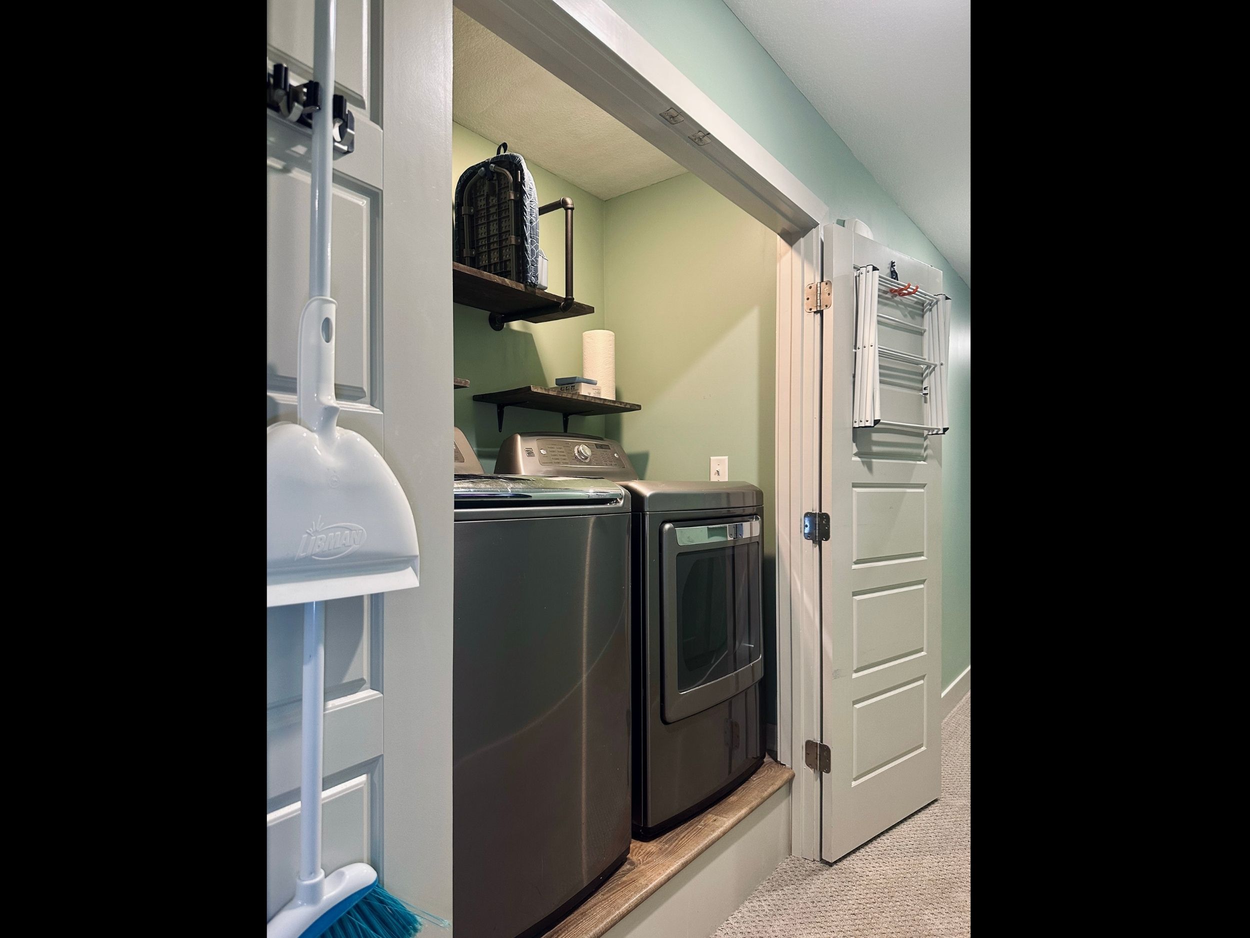 Convenient laundry closet featuring washer and dryer, ironing board, hanging rack, paper towels, and dryer sheets for everyday ease.