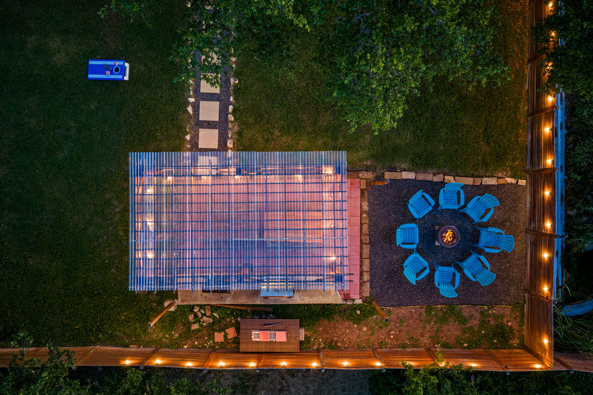 Aerial nighttime view of the backyard featuring a covered BBQ pavilion, glowing fire pit lounge, and spacious fenced yard. Designed for unforgettable evenings, outdoor dining, and relaxing under the Texas stars.