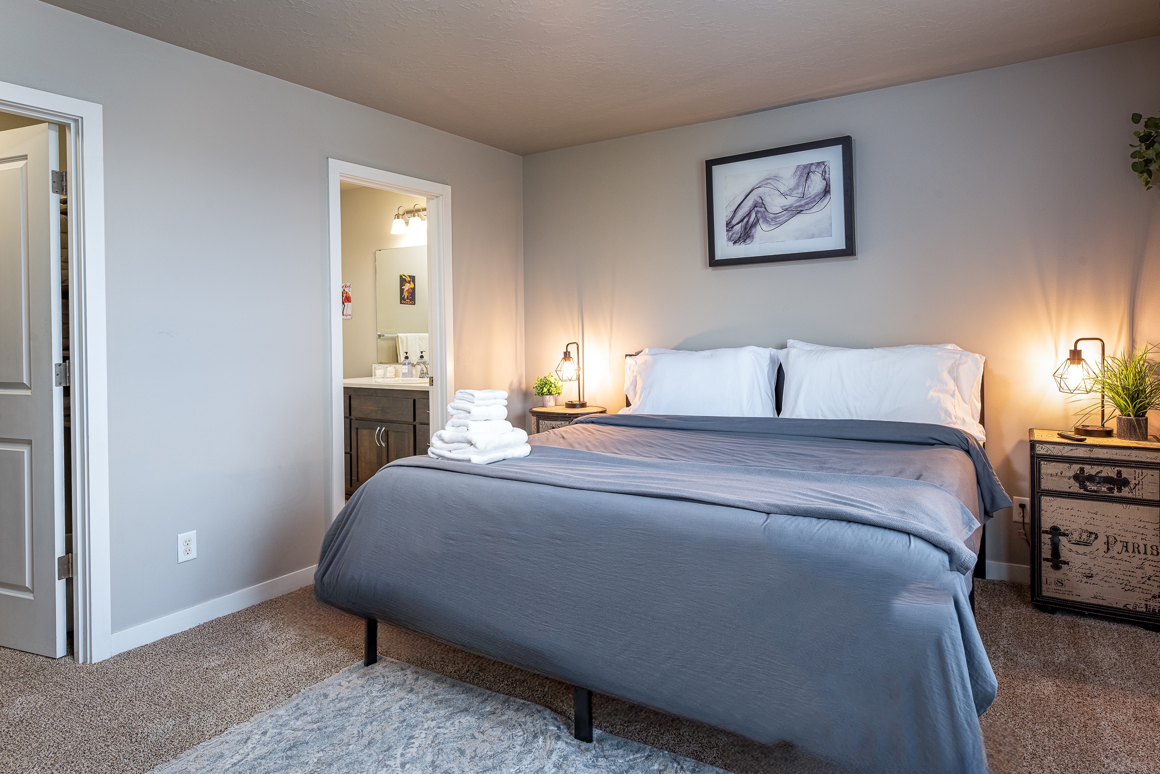 A cozy bedroom features a neatly made bed with a gray blanket and white pillows, a bedside table with a small plant and a decorative lamp, and a doorway leading to a bathroom with visible towels and toiletries.